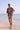 A man in a patterned shirt and shorts walking along the beach, with the ocean in the background.
