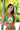 A woman in a green bikini holds a decorative fan with palm trees, standing in a tropical setting.