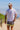 A man in a white t-shirt and colorful shorts stands on a sandy beach with the ocean in the background.