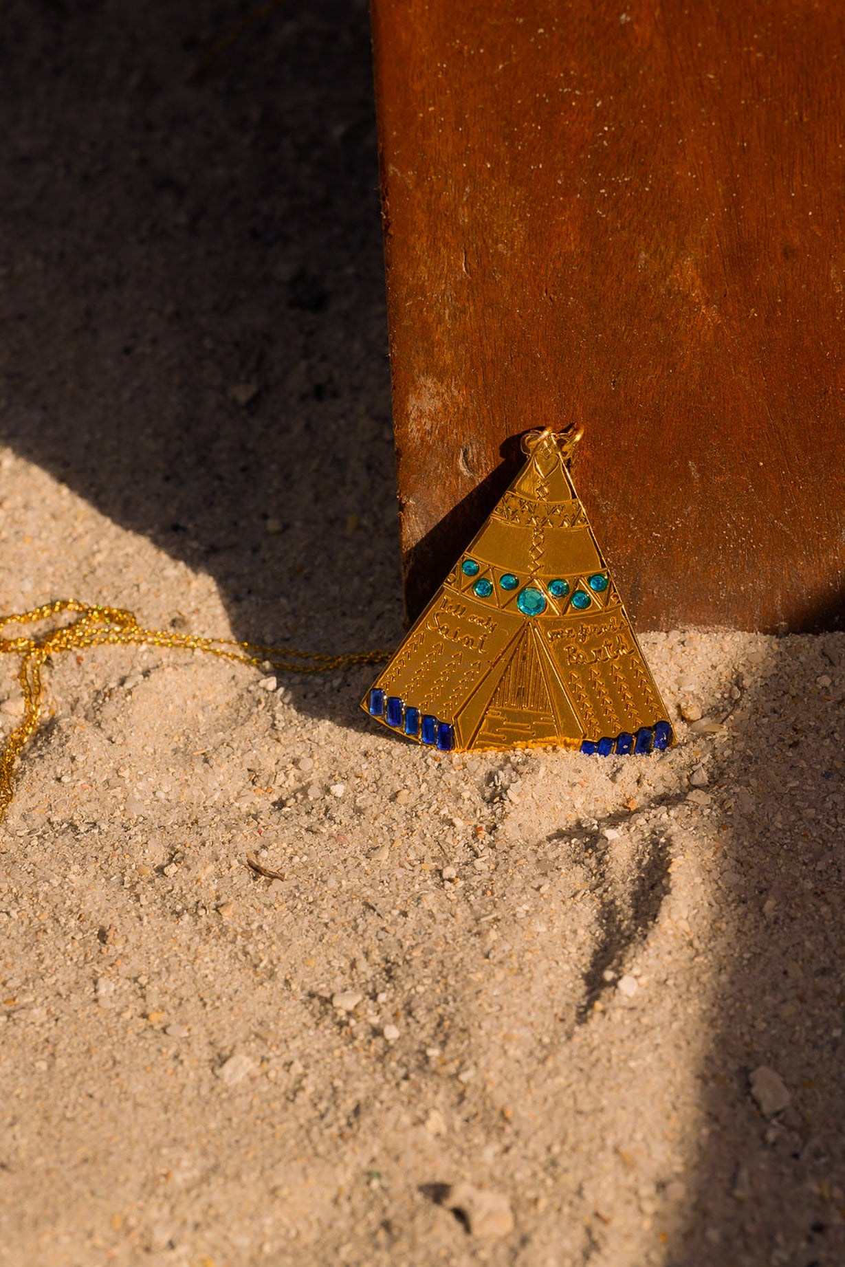 Gold triangular pendant with blue and green accents on sand near a wooden post.