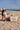 Person sitting on the beach with a hat and sunglasses, beside a large beach bag.